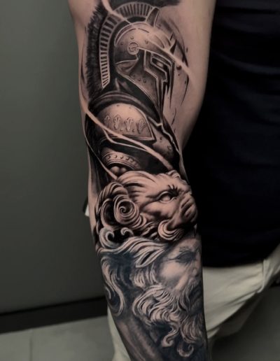 Tatuaje en estilo realista en el antebrazo de un guerrero espartano con un casco detallado, acompañado de una cabeza de león y un rostro de una divinidad clásica. La composición en tonos de negro y gris utiliza un sombreado impecable para aportar profundidad y textura, resaltando la fuerza y la majestuosidad del diseño.