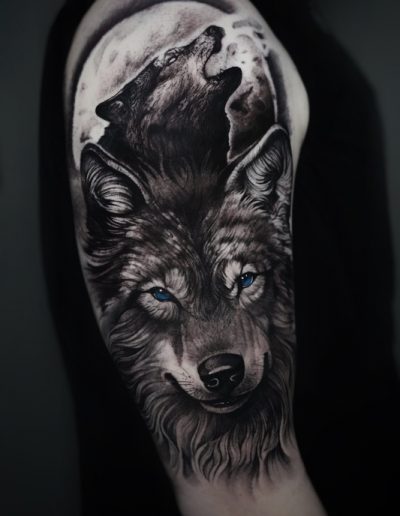 Tatuaje en estilo realista en el brazo de un lobo con mirada intensa y ojos azules, acompañado de la silueta de otro lobo aullando bajo una luna llena. El diseño en tonos de negro y gris resalta la textura del pelaje y los detalles de la luna, creando una imagen poderosa y mística.