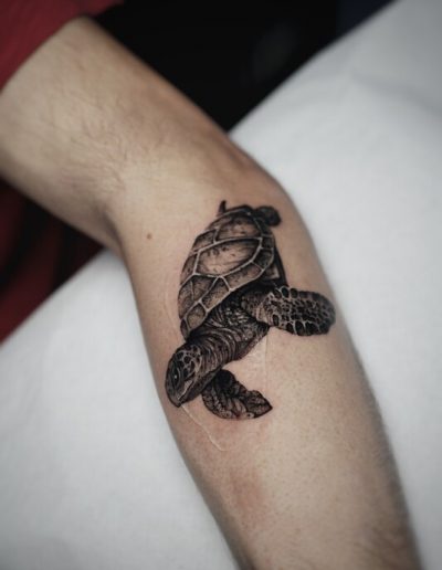 Tatuaje en estilo realista de una tortuga marina en tonos negro y gris, ubicado en el antebrazo de una persona. La tortuga parece estar nadando, con detalles intrincados en su caparazón y extremidades que resaltan su textura natural. La técnica de sombreado y el uso del contraste crean un efecto tridimensional, dando la impresión de que la tortuga emerge de la piel.