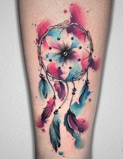 Tatuaje de un atrapasueños en estilo acuarela, con tonos azul, rosa y morado difuminados. El diseño presenta plumas colgantes y una flor en el centro del aro, creando un efecto etéreo y artístico con un fondo de manchas de color.