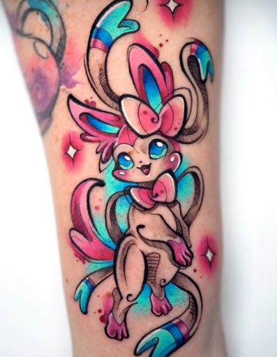 Tatuaje en estilo acuarela de una criatura mágica con orejas largas y lazos, con tonos vibrantes de azul, rosa y morado. El diseño incorpora detalles en negro que resaltan la figura sobre un fondo difuminado de manchas de color y destellos brillantes, creando un efecto dinámico y artístico.