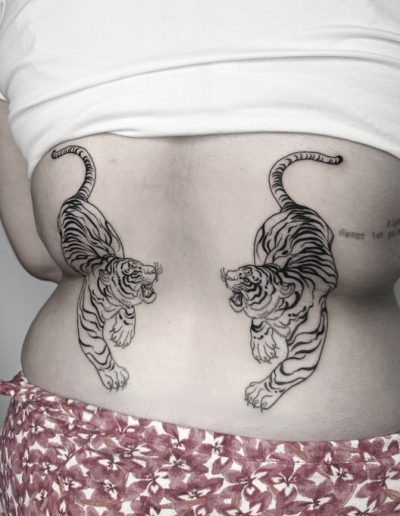 Tatuaje fineline en Barcelona con diseño simétrico de dos tigres en la zona lumbar. Arte detallado con trazos limpios y estilo minimalista.